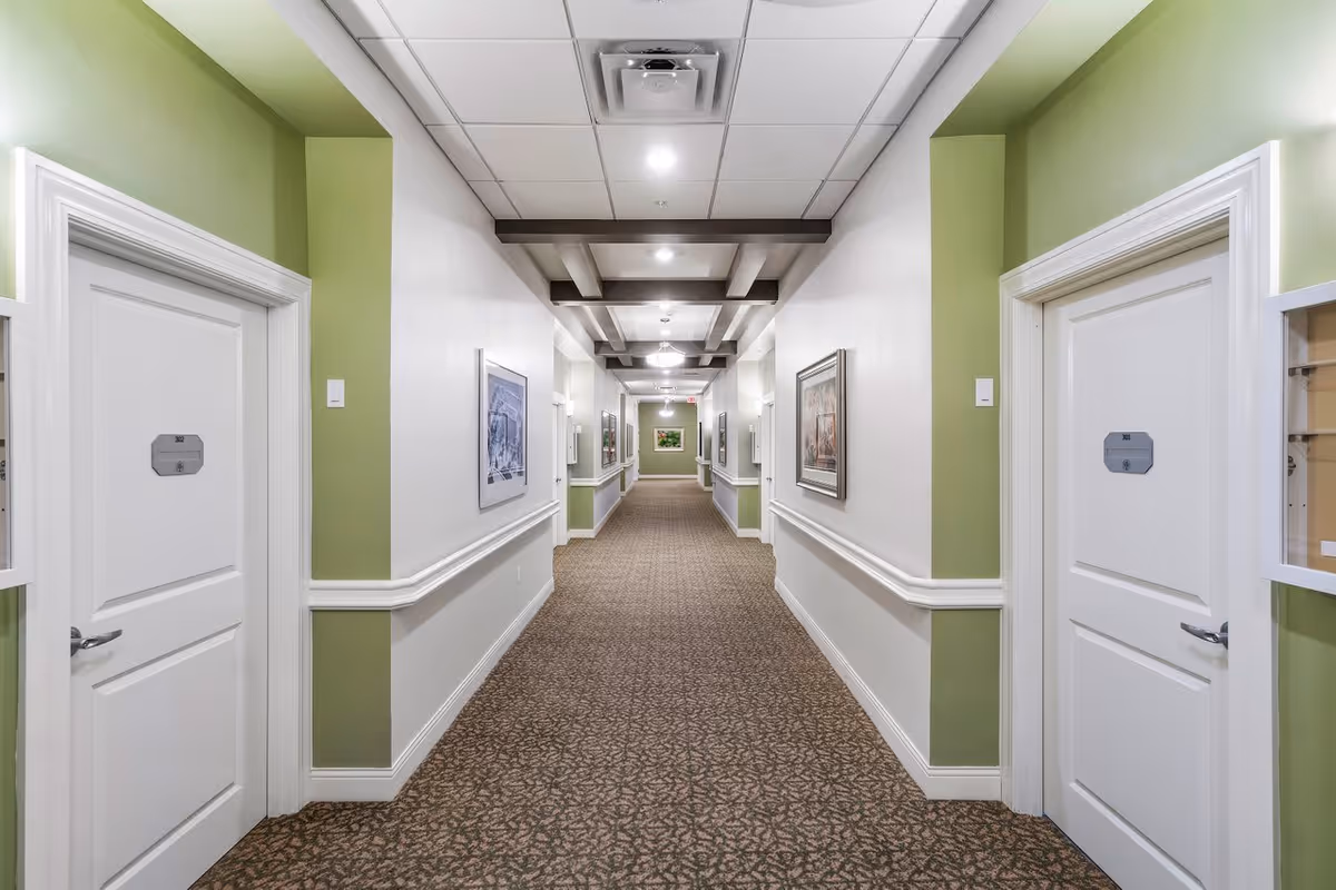 A long, well-lit hallway in a senior living facility with white doors on either side, green and white walls, framed artwork, and a patterned carpet. The ceiling has recessed lighting and decorative beams.