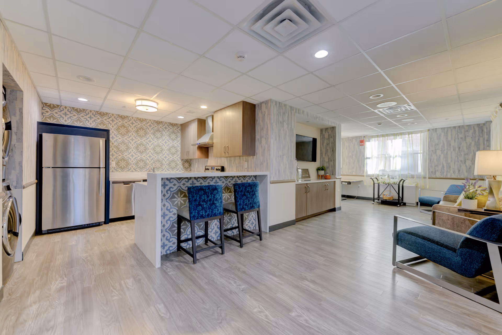A bright and modern interior space featuring a kitchen area with stainless steel refrigerator, dishwasher, stove, and wooden cabinets. The kitchen island has a decorative tile front and two blue upholstered bar stools. Adjacent to the kitchen is a living area with blue and beige chairs, a side table with a lamp, a wall-mounted TV, and a window with sheer curtains allowing natural light to fill the room. The flooring is light wood, and the ceiling has recessed lighting and a central light fixture.