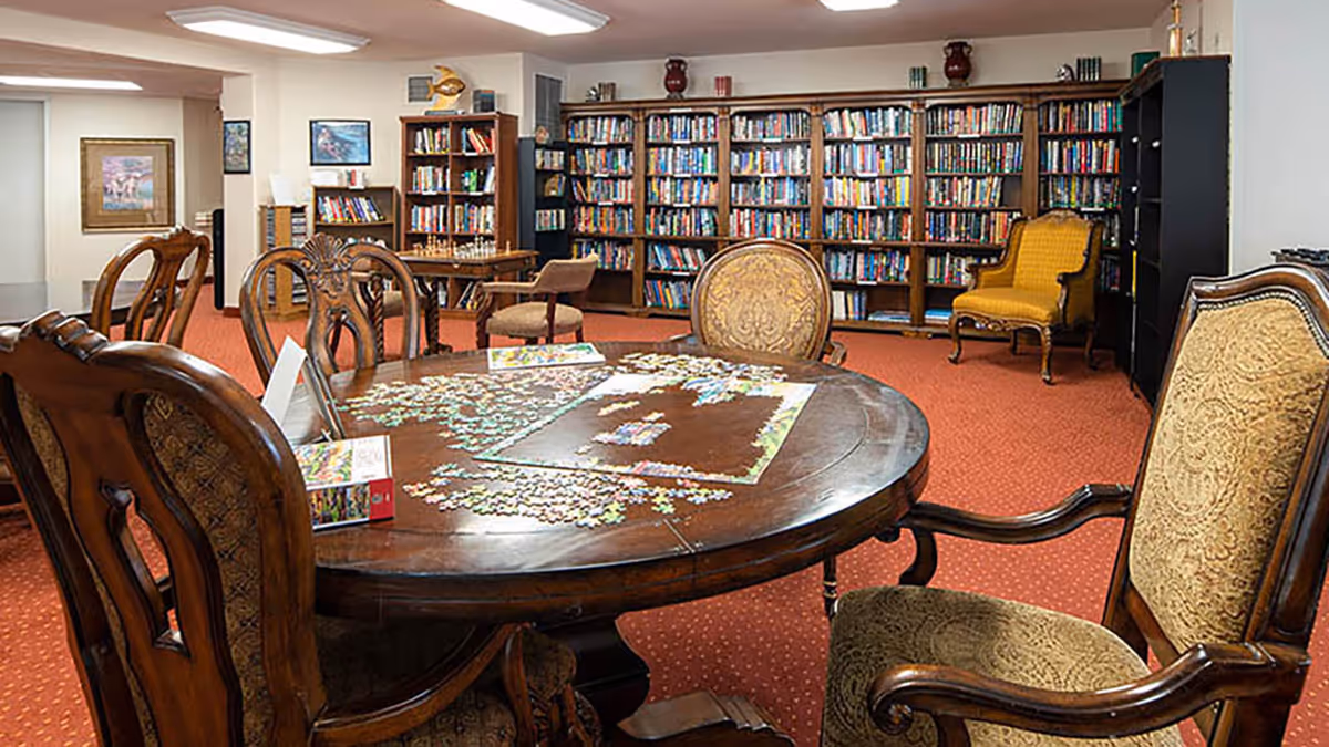 A cozy library/activity room with a round wooden table and chairs holding a partially completed jigsaw puzzle, and bookshelves along the back wall.