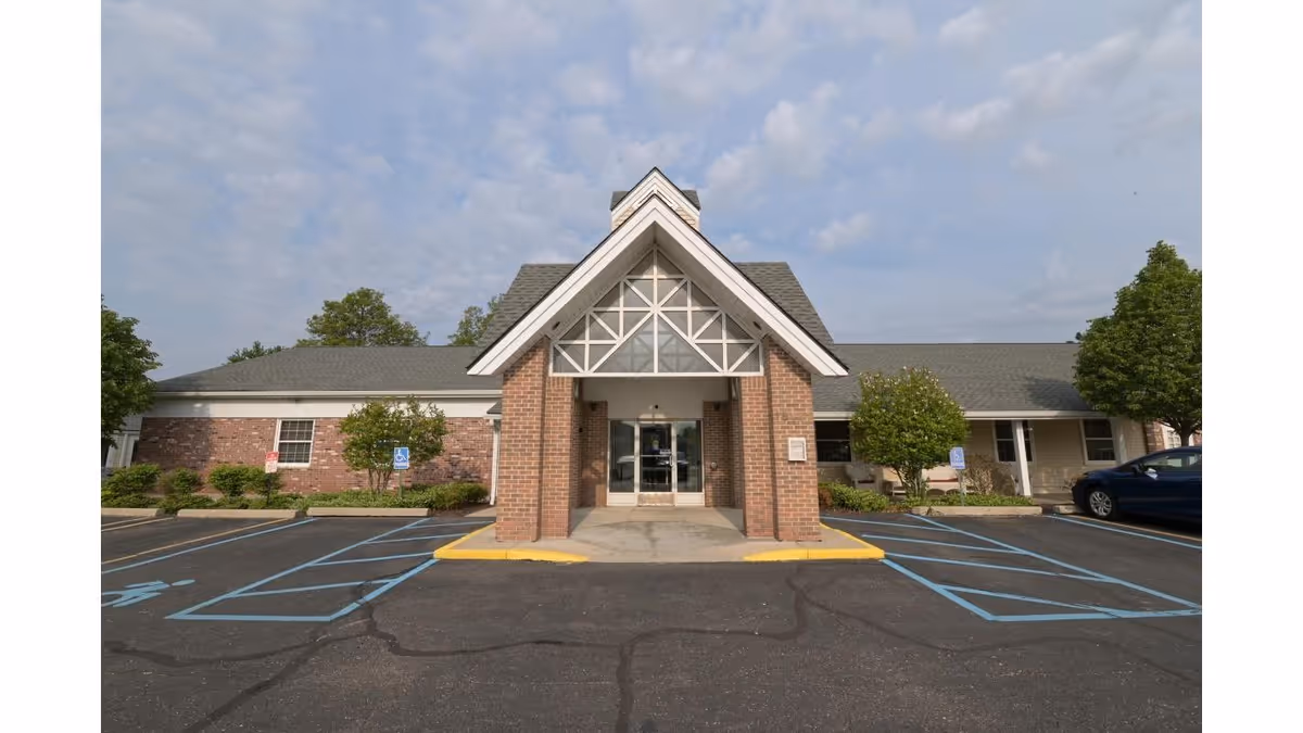 Front exterior view of a single-story brick building with a peaked roof and a covered entrance. There are handicap parking spaces marked in blue on the asphalt parking lot in front of the building. Trees and shrubs are planted along the building's facade under a partly cloudy sky.
