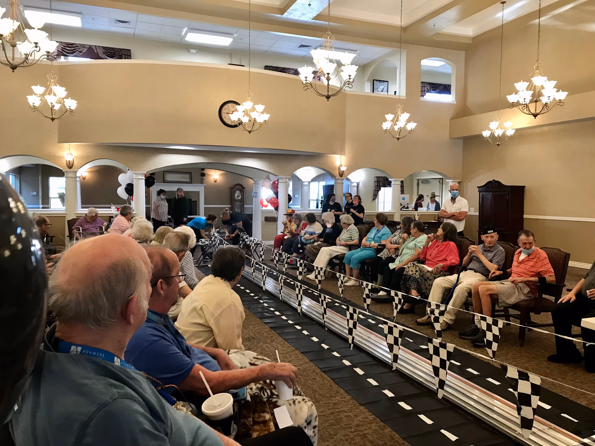 A large group of elderly people seated in chairs along both sides of a checkered flag decorated race track inside a spacious, well-lit common area with chandeliers and beige walls. Some staff members are standing in the background, and a few people are wearing face masks.