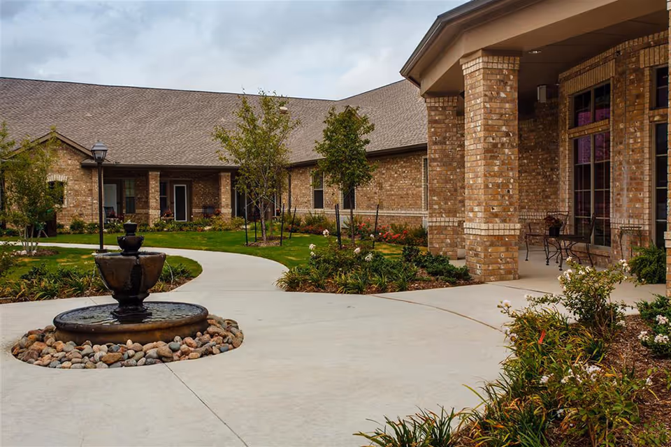 Outdoor courtyard area of The Harrison At Heritage facility featuring a circular water fountain surrounded by rocks, a curved concrete walkway, landscaped garden beds with flowers and shrubs, young trees, and a brick building with large windows and a covered porch with metal chairs and tables.