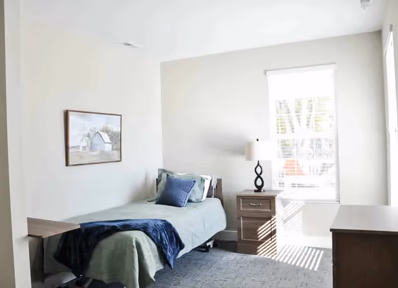 A bright bedroom with a single bed covered in green bedding and a blue throw blanket, a wooden nightstand with a lamp, a framed picture on the wall, and a window letting in natural light.
