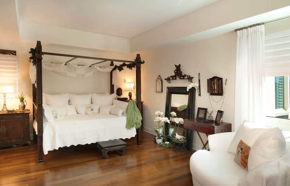 Sunlit bedroom with a dark wood four-poster bed dressed in white linens, wooden floors, and a seating area by the window.