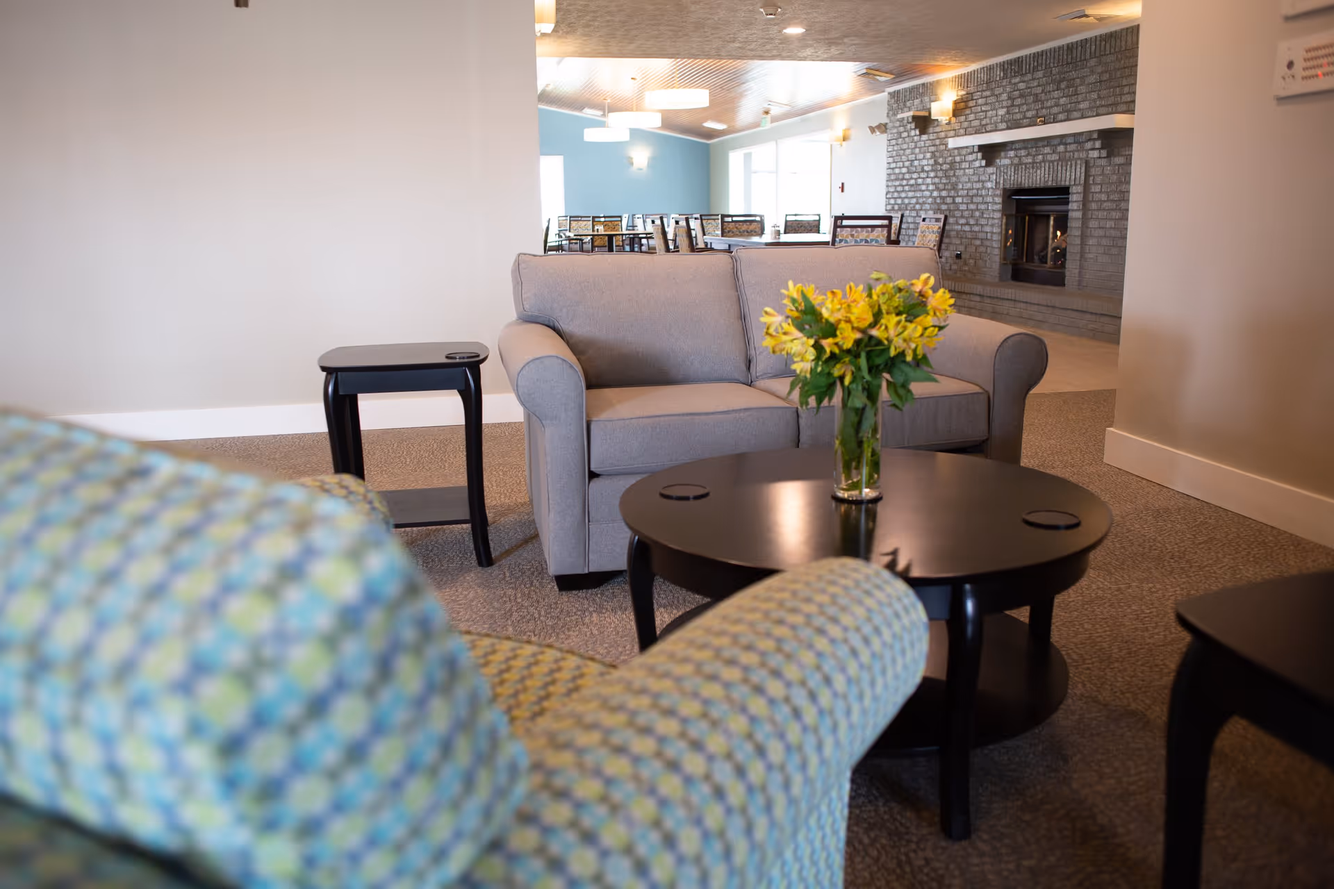 A cozy living room area with a gray loveseat, a patterned armchair, and a round black coffee table with a vase of yellow flowers. In the background, there is a brick fireplace and a dining area with tables and chairs.