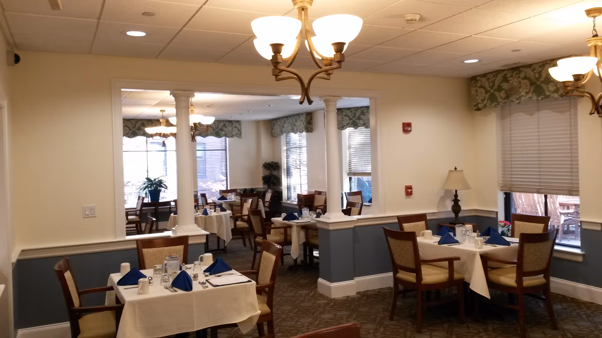 A dining room in an assisted living facility with several tables covered with white tablecloths, each set with blue folded napkins, cups, glasses, and silverware. The room has beige walls with blue wainscoting, carpeted floors, and multiple windows with floral valances. There are chandeliers hanging from the ceiling providing warm lighting, and a small table lamp is visible near one window.