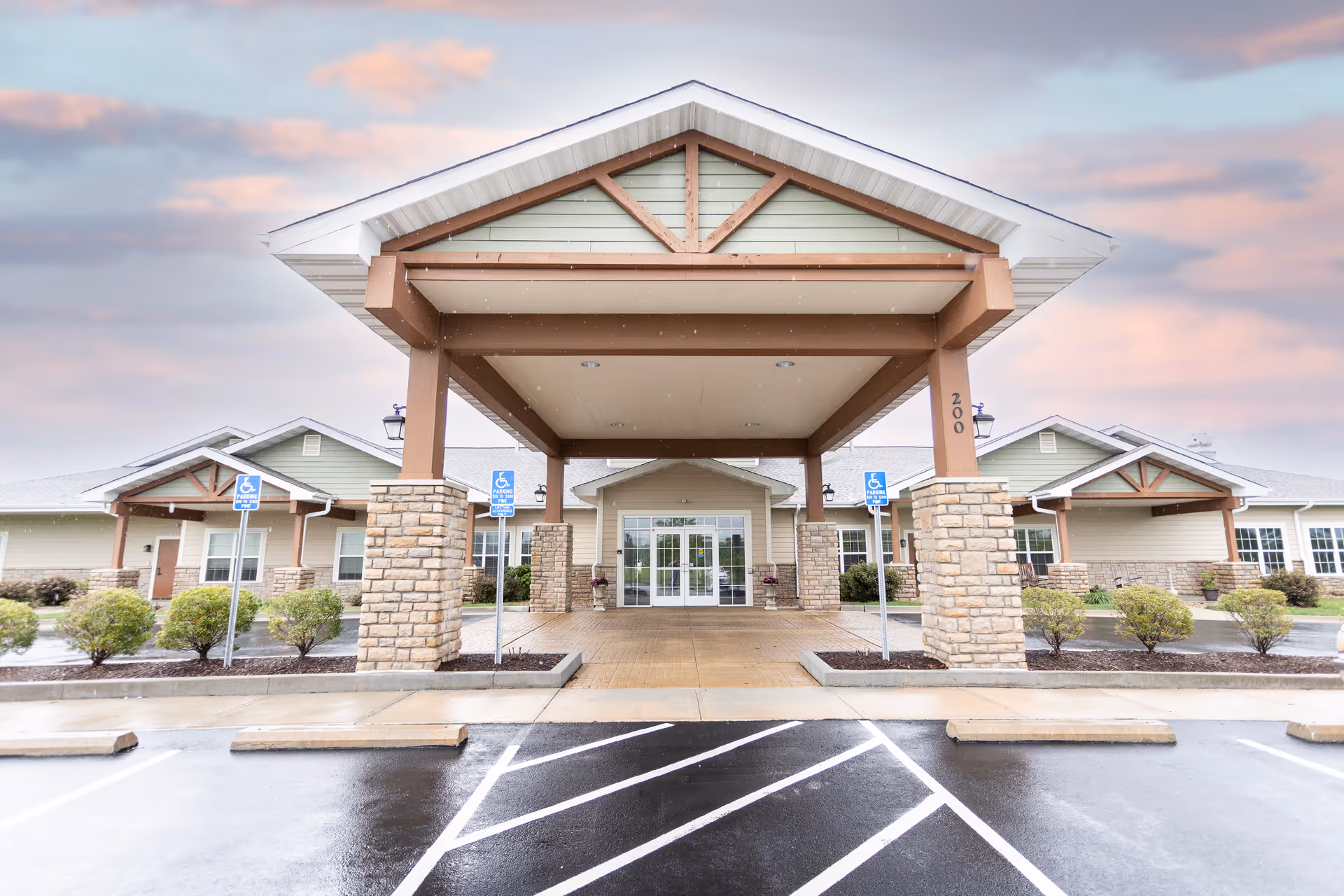 Front exterior view of Oak Pointe of Kearney facility showing a covered entrance with stone pillars, handicap parking spaces, and a cloudy sky at sunset.