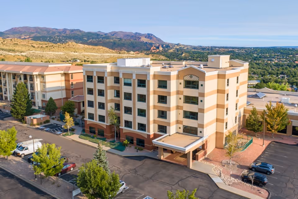 Aerial view of a multi-story senior living facility building with a covered entrance, surrounded by parking lots and trees, with mountains visible in the background under a clear sky.