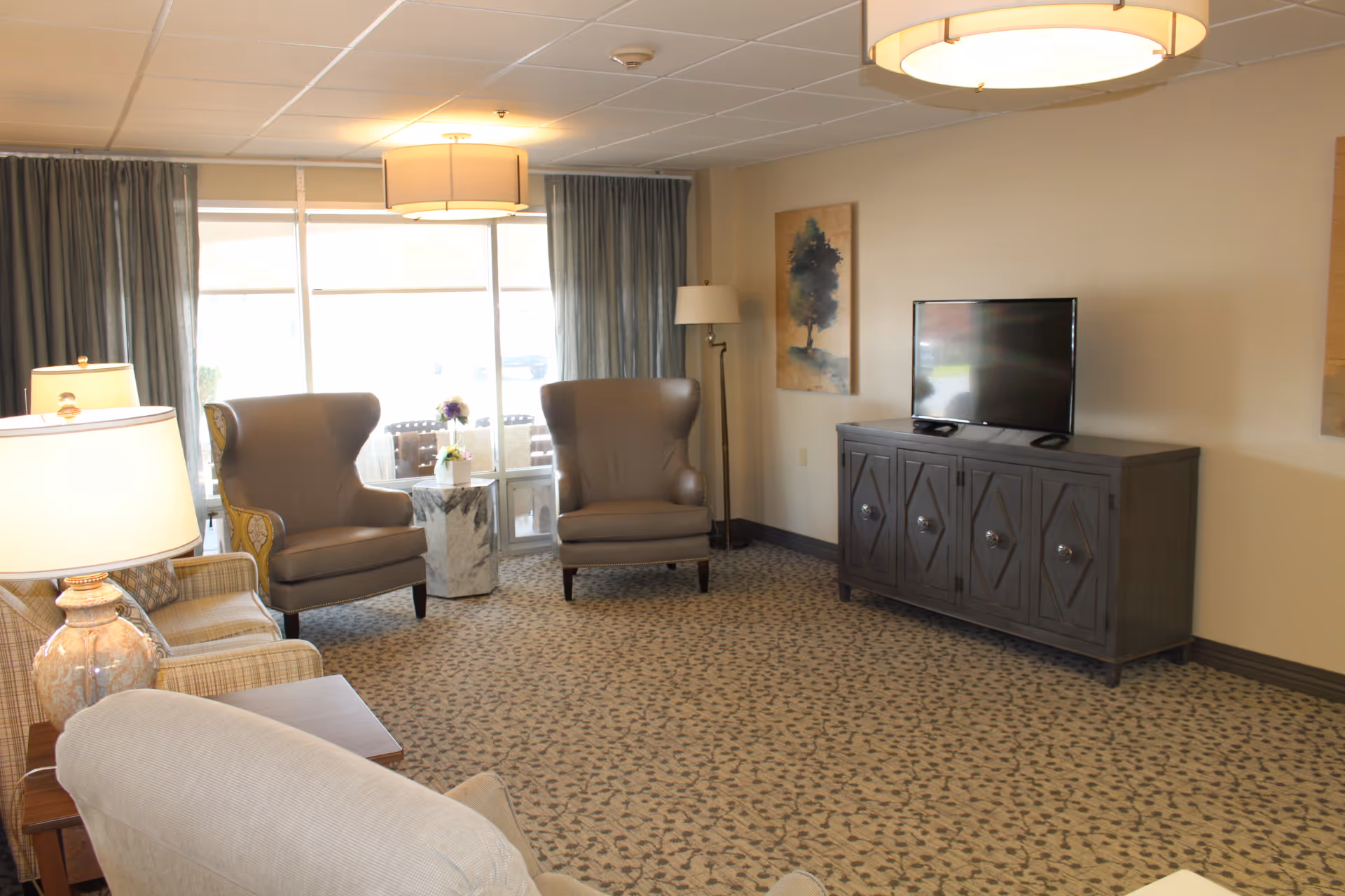 A cozy living room with patterned carpet, two brown armchairs, a beige sofa, a wooden TV cabinet with a flat-screen TV, two table lamps, a floor lamp, and a large window with curtains letting in natural light.