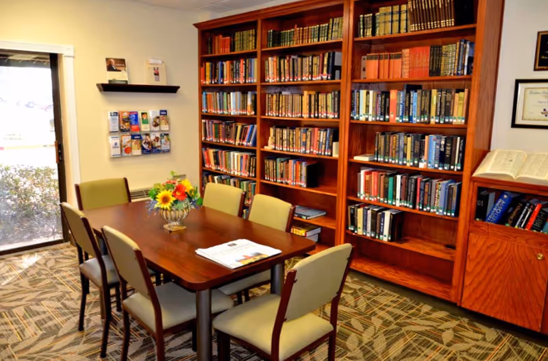 Small common room with a wooden table and chairs, a floral centerpiece, and large bookshelves filled with books.