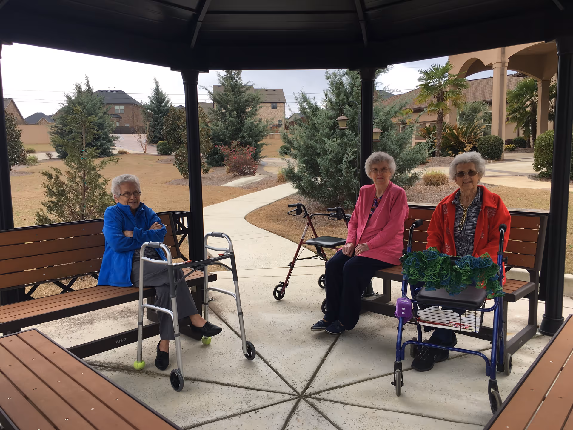 Three elderly women sitting on benches under a gazebo in an outdoor garden area. Two of the women have walkers in front of them. The background shows trees, shrubs, and a pathway with residential buildings visible in the distance.