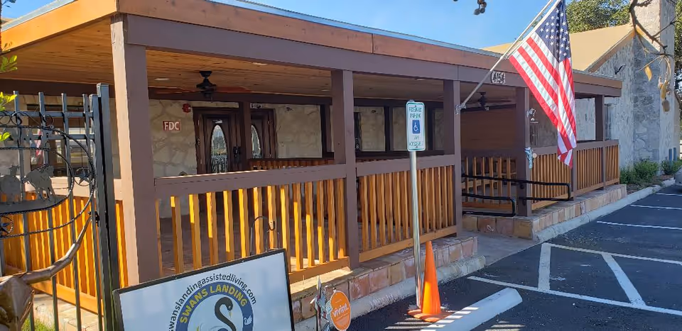 Exterior view of Swans Landing Assisted Living facility showing a covered wooden porch with railings, an American flag mounted on a pole, a handicap parking sign, and a parking lot with marked spaces. The building has stone walls and wooden doors.