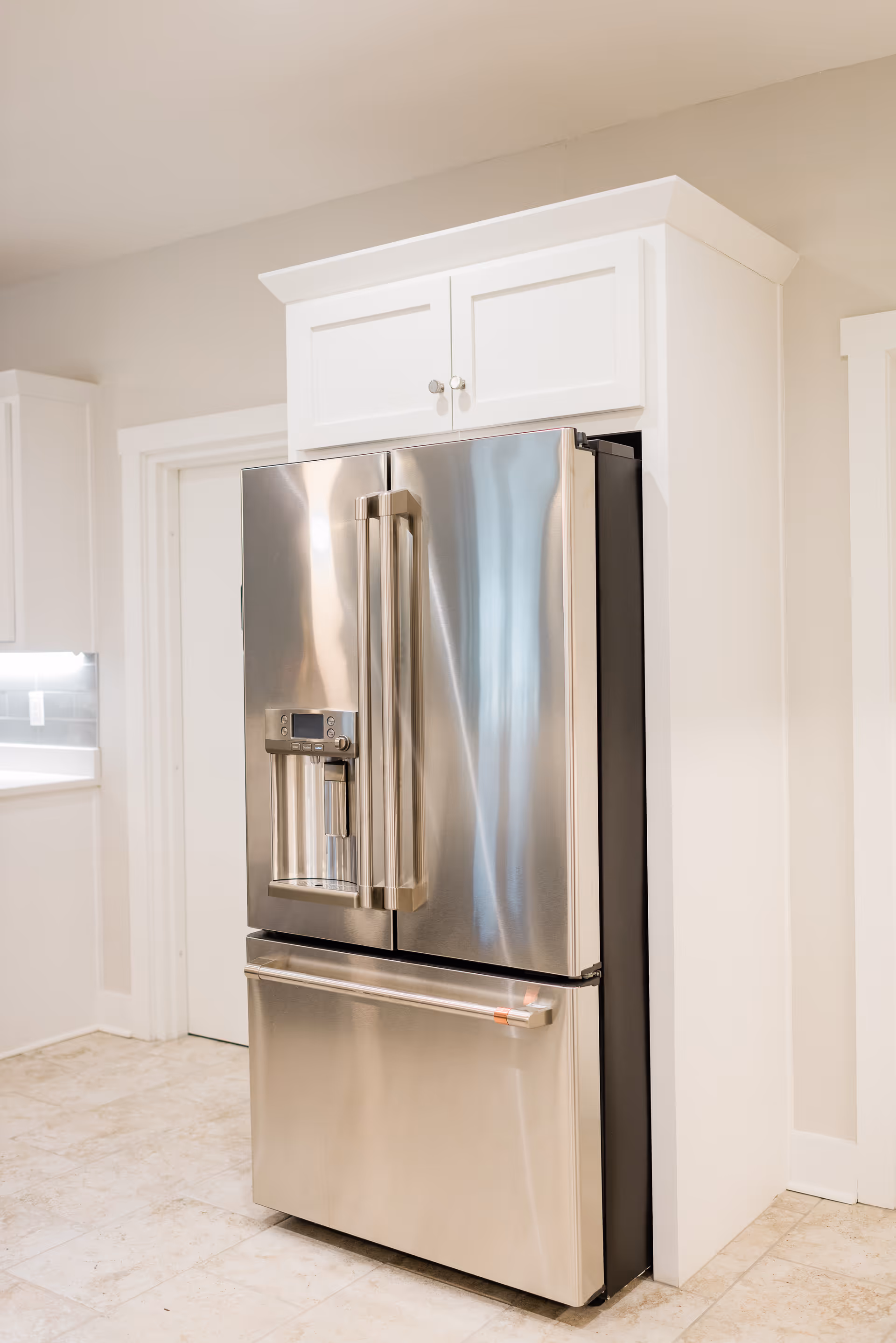A modern stainless steel French door refrigerator with a bottom freezer drawer, set within white cabinetry in a bright kitchen with light-colored tile flooring.