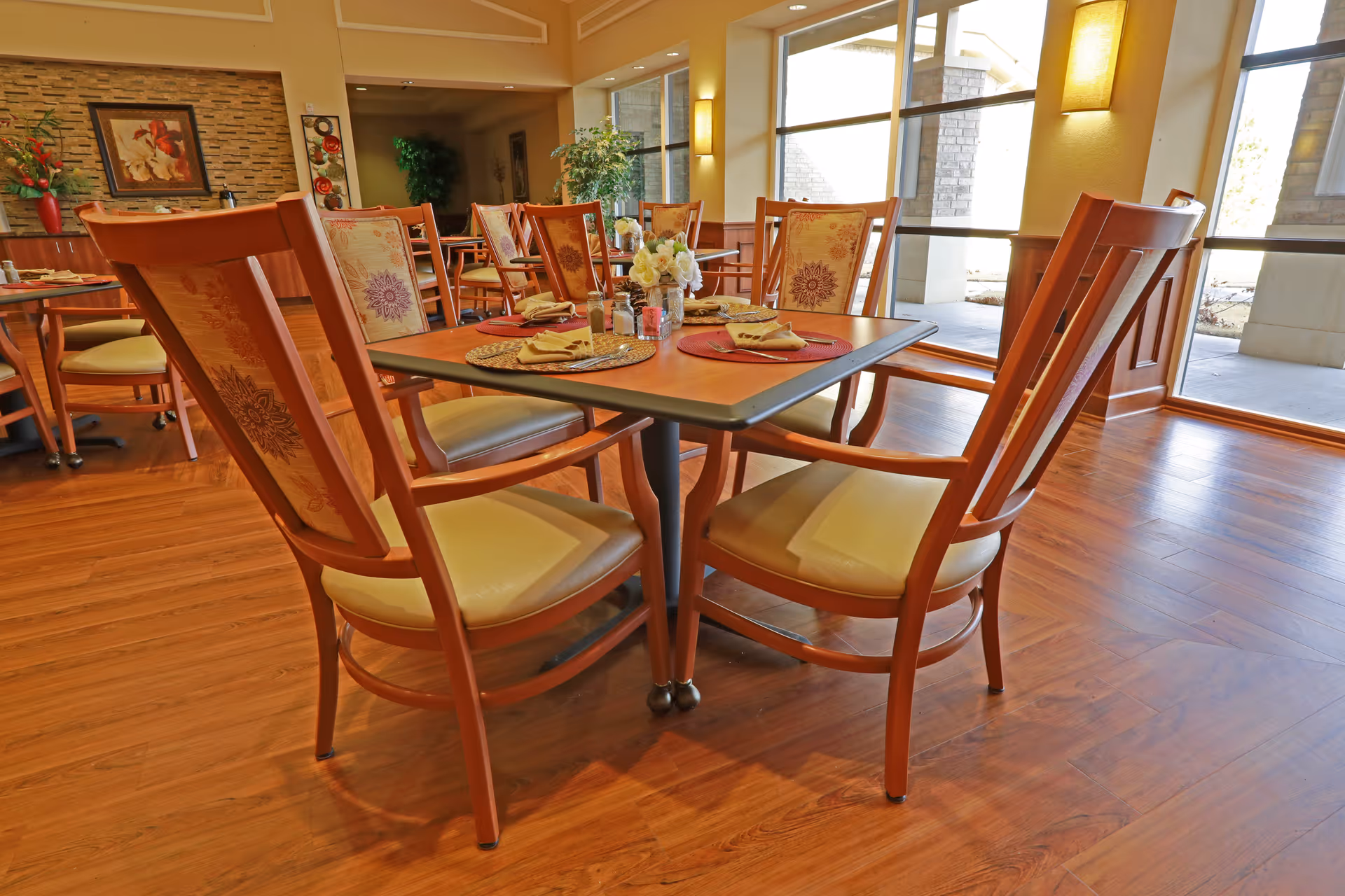 Well-lit dining area with wooden tables and chairs set with placemats and napkins next to large windows and hardwood floors.