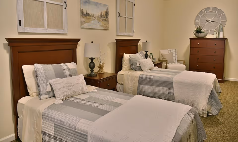 Two twin beds with gray plaid bedding in a softly lit, well-decorated bedroom with wooden headboards, a nightstand, and a dresser.