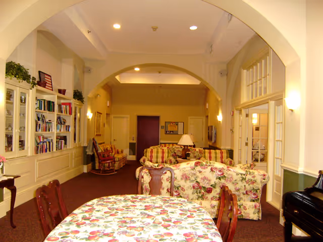 A warm communal lounge with floral upholstered sofas, a dining table in the foreground, bookcases along the left wall, and arched ceilings.