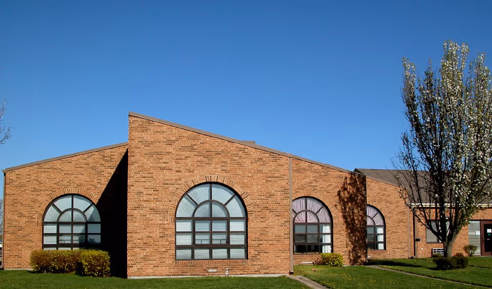 Brick single-story building facade with large arched windows, lawn and trees under a clear blue sky.