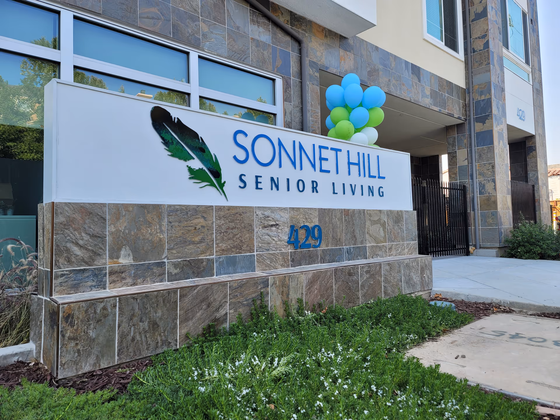 Exterior view of the entrance sign for Sonnet Hill Senior Living, featuring a large white sign with a feather logo and blue text, surrounded by stone tiles and greenery, with blue and green balloons attached near the entrance.