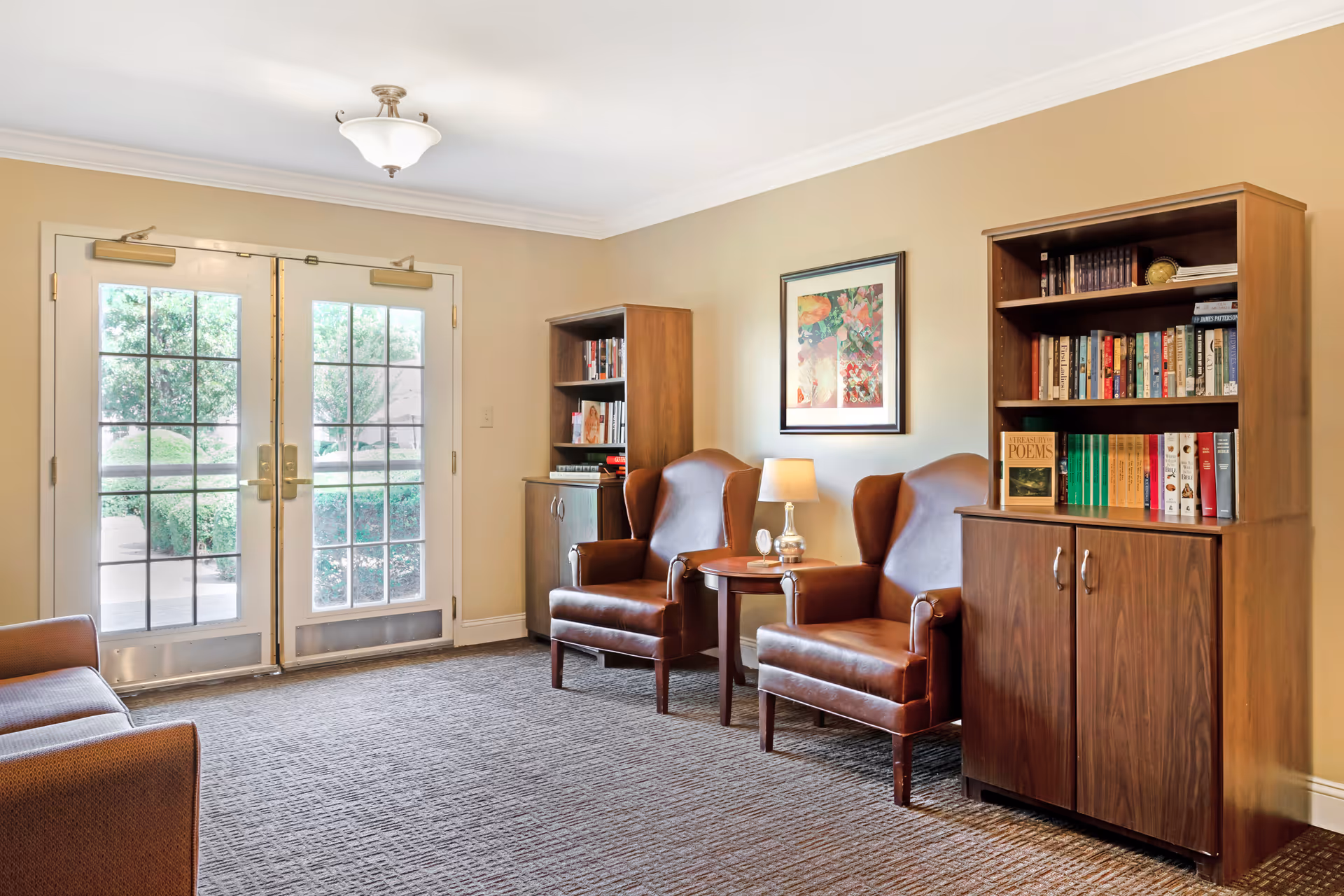 A cozy sitting area in a senior living facility with two brown leather armchairs and a small round wooden table with a lamp between them. There are two wooden bookshelves filled with books on either side of the chairs. A framed floral artwork hangs on the beige wall above the chairs. Double glass doors with a view of greenery outside are on the left side of the room. The floor is carpeted in a neutral tone.