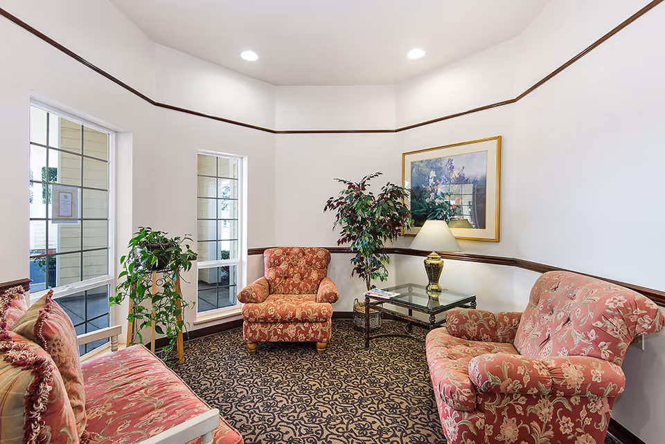 A cozy sitting area in a retirement community with two upholstered armchairs and a matching loveseat in a red floral pattern. There is a glass-top side table with a decorative lamp and a framed floral painting on the wall. Two large windows let in natural light, and there are two potted plants adding greenery to the space. The carpet has a dark intricate pattern, and the walls are white with a dark wood trim.