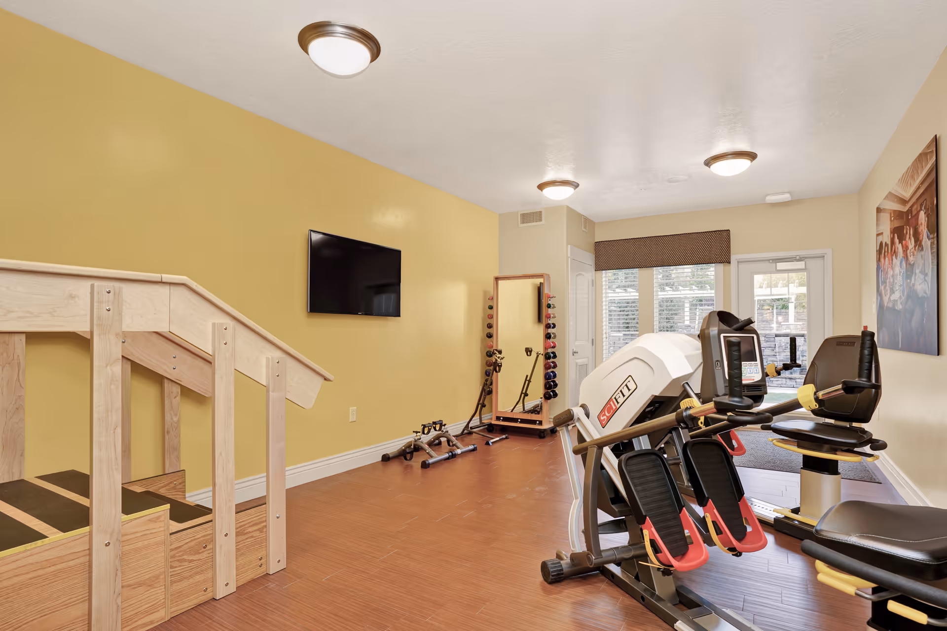 A small fitness room with light yellow walls and wood flooring. The room contains exercise equipment including a SciFit machine, a recumbent bike, a set of dumbbells on a rack, a full-length mirror, and a wooden staircase with handrails. A flat-screen TV is mounted on the wall, and there is a door and window with blinds letting in natural light.