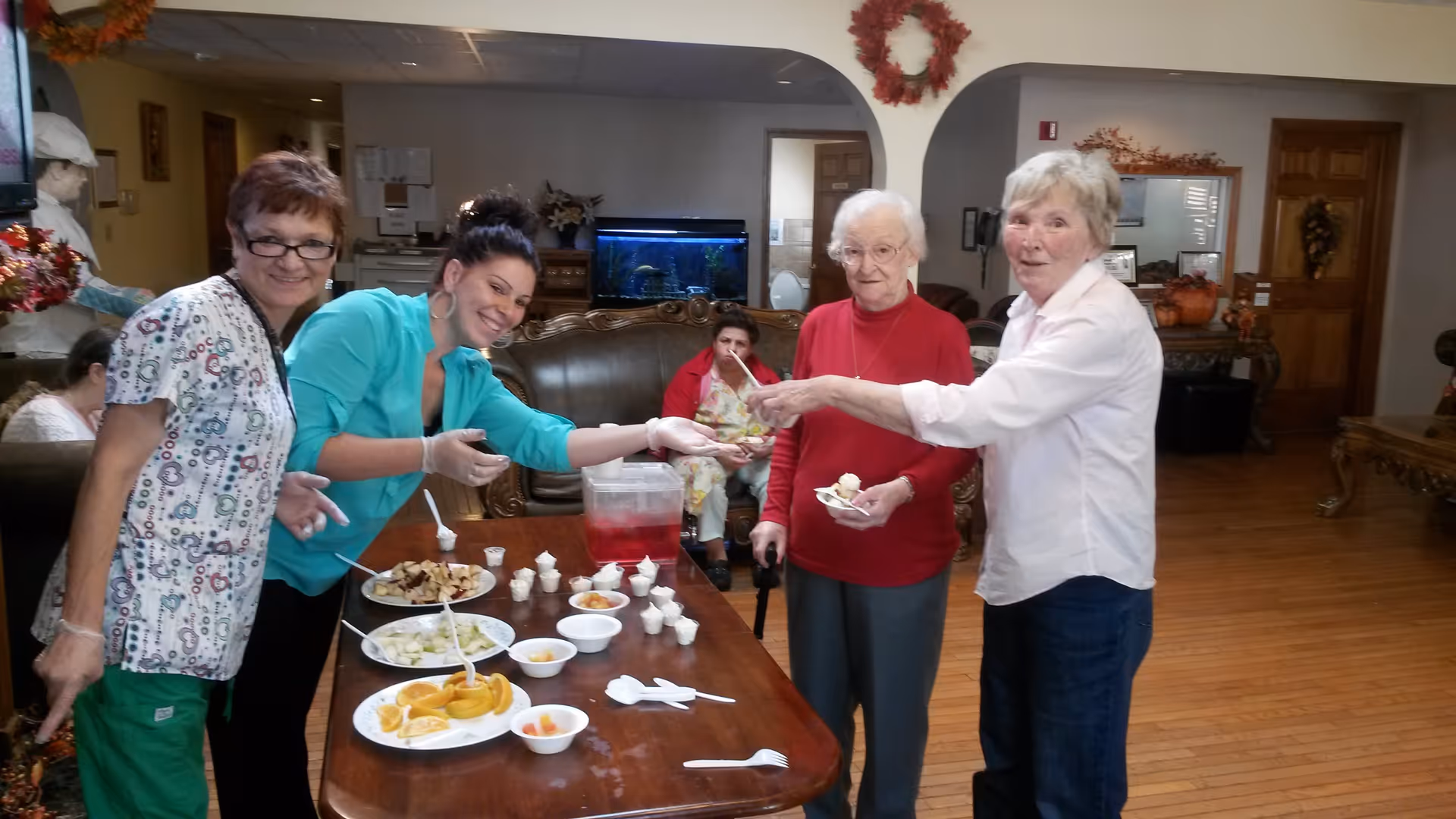 A group of elderly women and a caregiver are gathered around a wooden table with plates of food and small cups. The caregiver is serving food to one of the elderly women. The room has wooden floors, a fish tank, and autumn-themed decorations on the walls.
