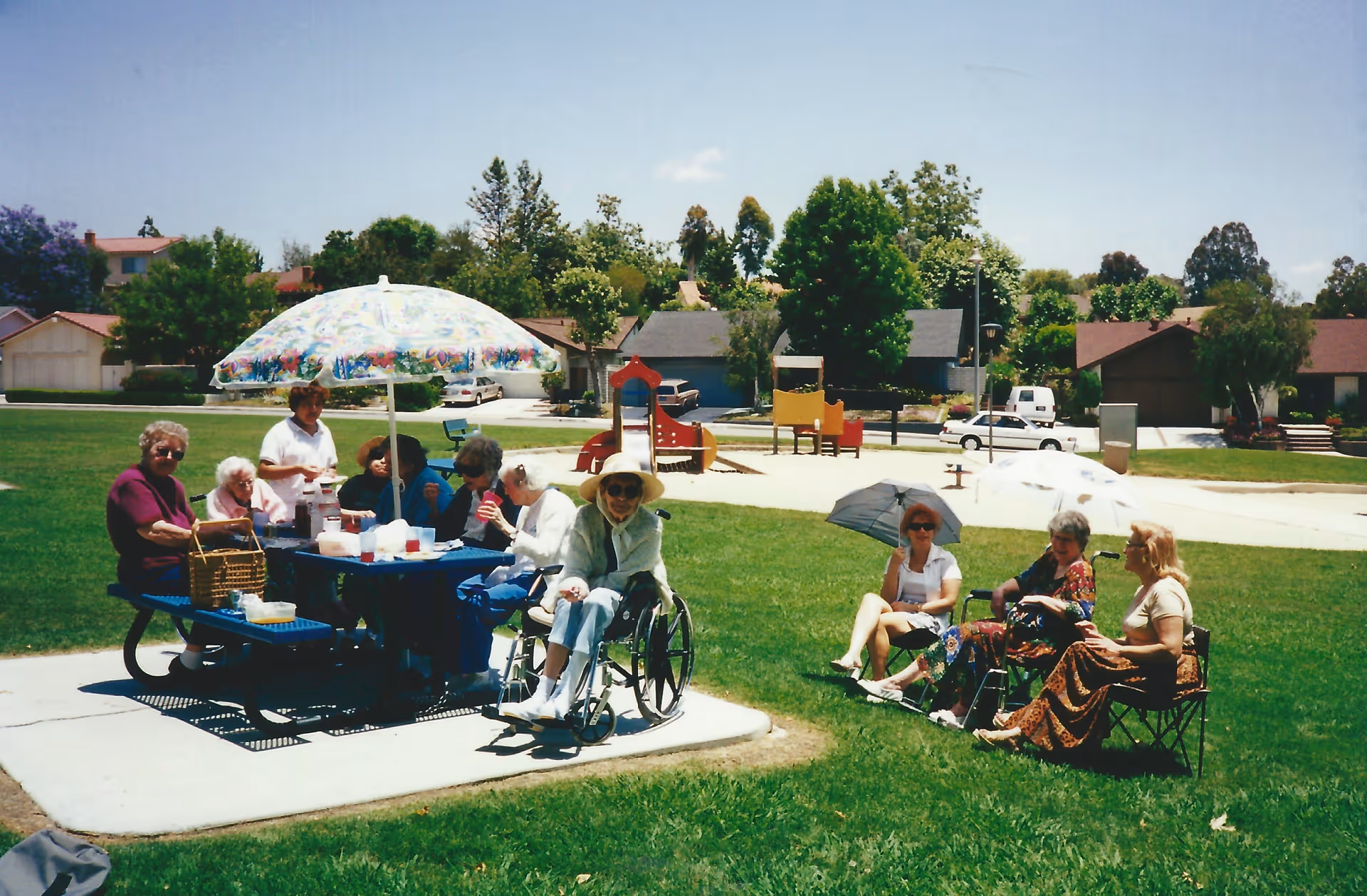 A group of elderly people gathered outdoors in a park-like setting. Some are seated at a picnic table under a colorful umbrella, while others sit on chairs on the grass holding parasols. There is playground equipment and houses in the background under a clear blue sky.