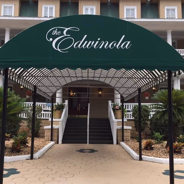 Entrance to a building with a green canopy that reads 'the Edwinola' in white cursive letters. The canopy covers a walkway leading to stairs with white railings, flanked by plants and landscaping.