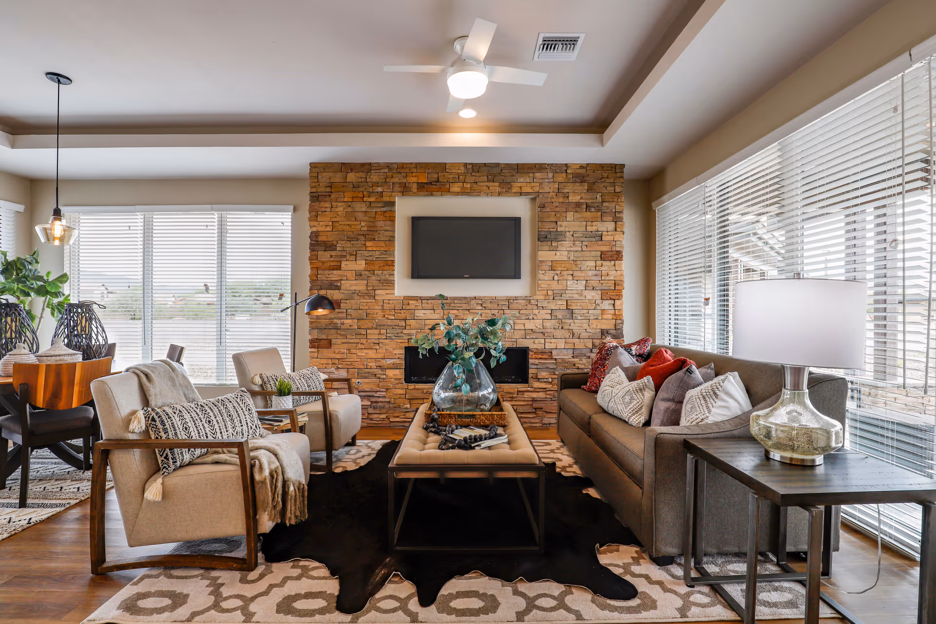 A cozy living room with a stone accent wall featuring a mounted flat-screen TV above a modern fireplace. The room has a gray sofa with multiple decorative pillows, two beige armchairs with patterned cushions and throws, a rectangular coffee table with a glass vase and books, a side table with a glass lamp, and large windows with white blinds letting in natural light. A dining area with a wooden table and chairs is visible in the background.