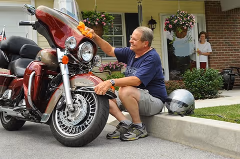 An elderly man is sitting on a curb outside a residential building, polishing a red motorcycle with an orange cloth. A woman stands in the doorway of the building, watching him. The building has hanging flower baskets and a well-maintained lawn.