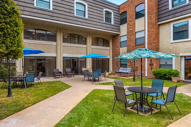 Outdoor courtyard area of a senior living facility with green grass, paved walkways, several tables with umbrellas, chairs, and a swing bench. The building surrounding the courtyard has multiple windows and a mix of beige and brick exterior walls.
