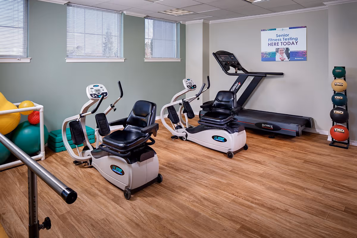 A senior fitness room with two recumbent exercise bikes, a treadmill, a rack of colorful medicine balls, and various exercise balls and steps in the corner. The room has wooden flooring, light green and white walls, and three windows with blinds. A sign on the wall reads 'Senior Fitness Testing HERE TODAY.'