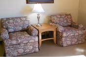 Two floral armchairs flank a wooden side table with a lamp between them.