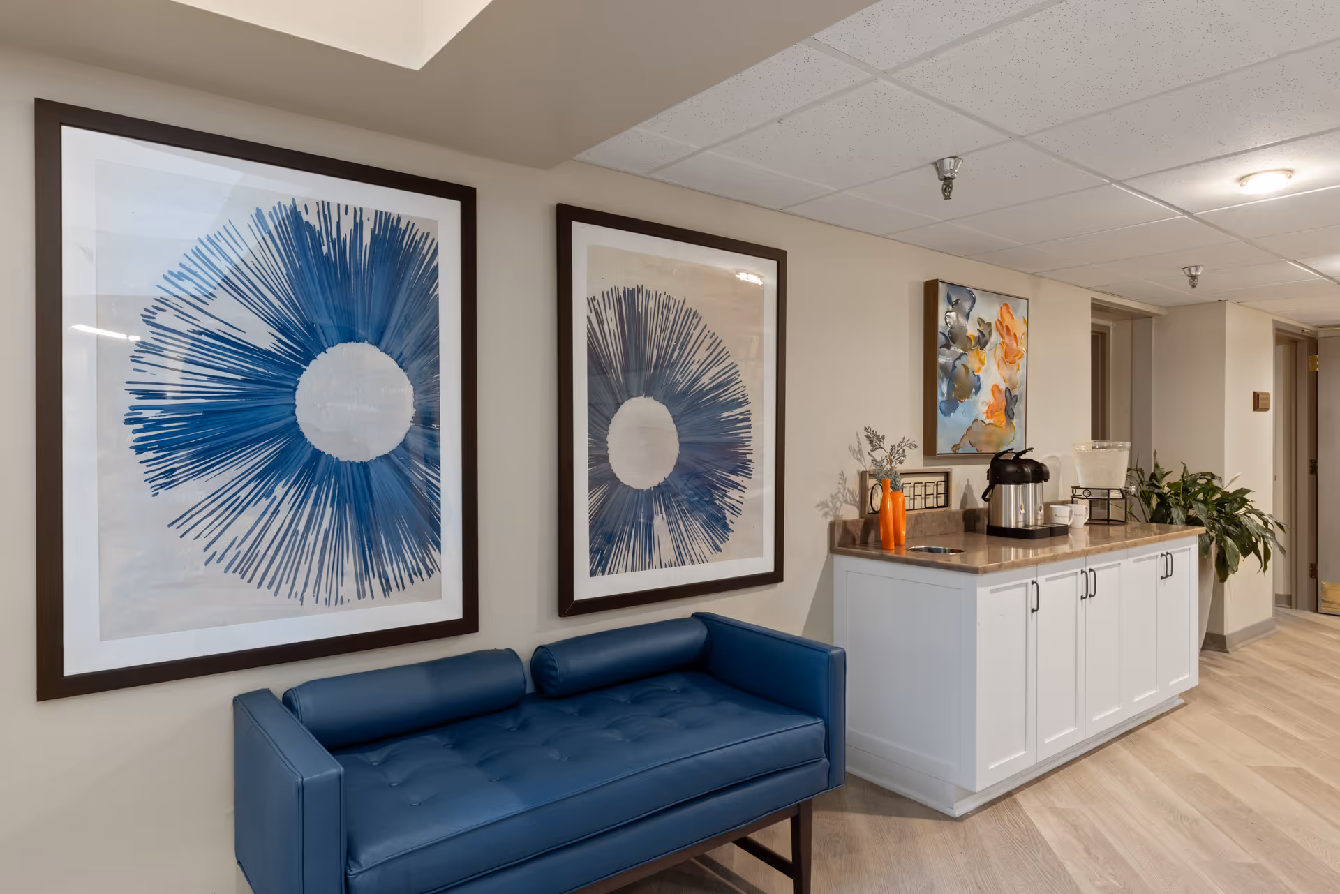 Interior seating area with a blue upholstered bench, two large blue abstract wall artworks, and a white cabinet countertop with a coffee station.