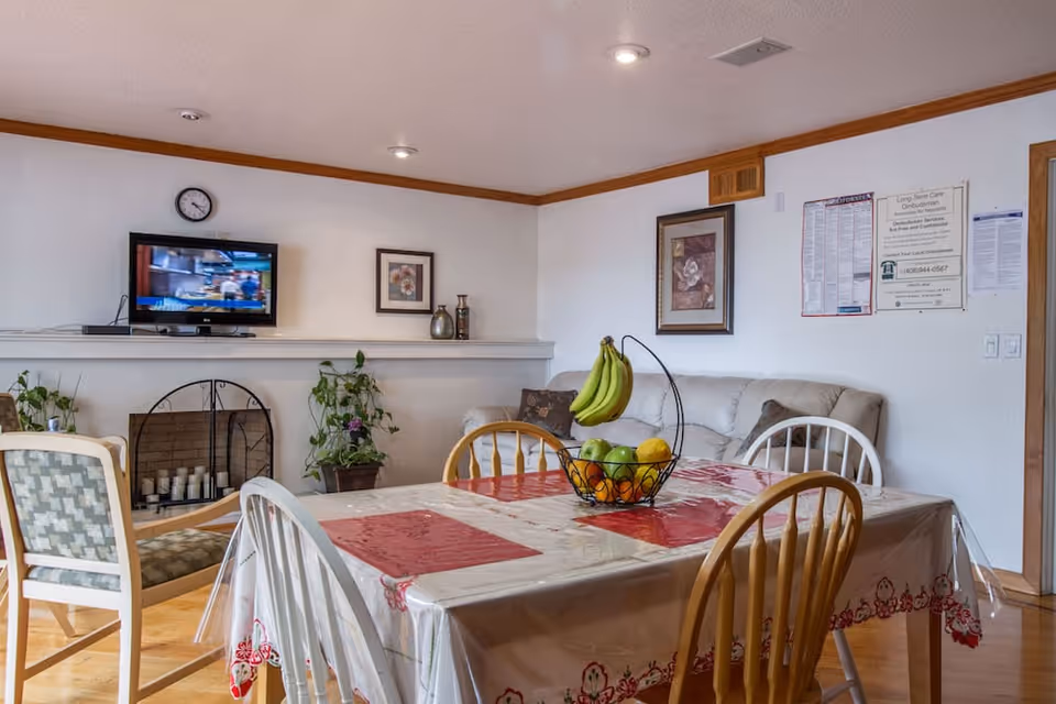 A cozy common area with a dining table covered with a plastic tablecloth and red placemats. The table has a fruit basket with bananas, apples, and oranges. Surrounding the table are wooden and cushioned chairs. In the background, there is a white couch with pillows, framed artwork on the walls, a small TV mounted above a fireplace with candles inside, and some potted plants.