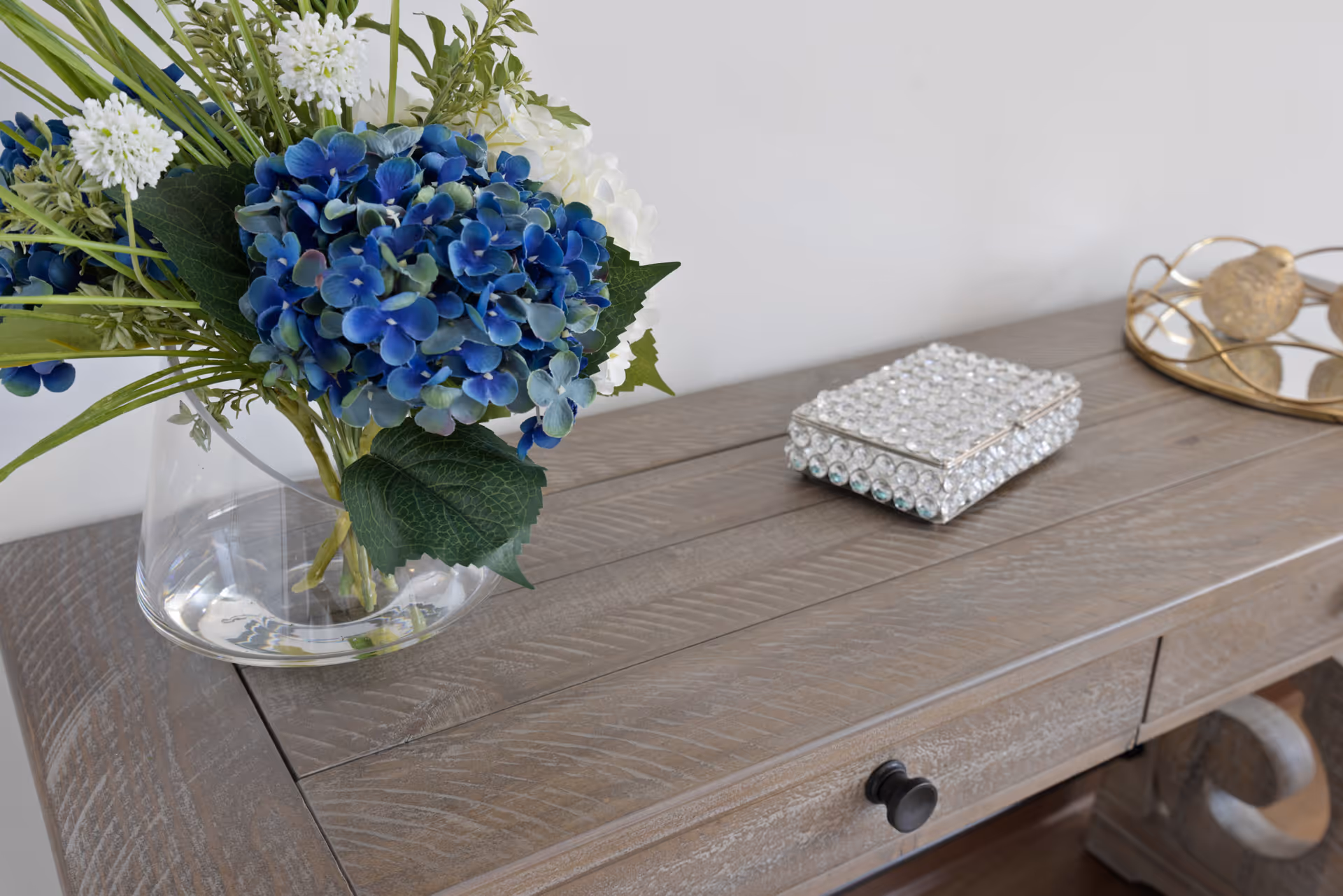 A wooden console table topped with a glass vase of blue and white flowers and decorative boxes.