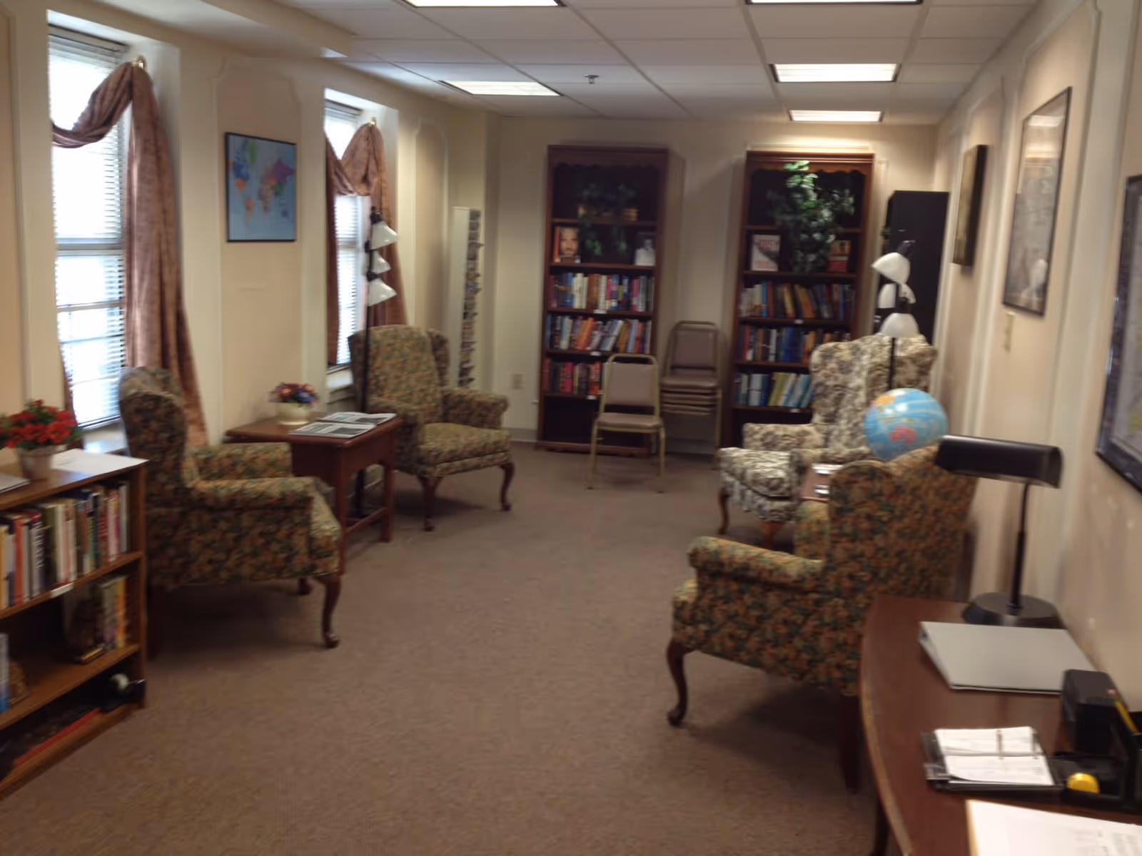 A cozy reading or sitting room with floral upholstered armchairs arranged around small wooden tables. There are two tall bookshelves filled with books and plants against the far wall. The room has beige walls, carpeted floor, and windows with pinkish curtains letting in natural light. A globe sits on one of the chairs, and a desk with a lamp, laptop, and papers is visible on the right side.