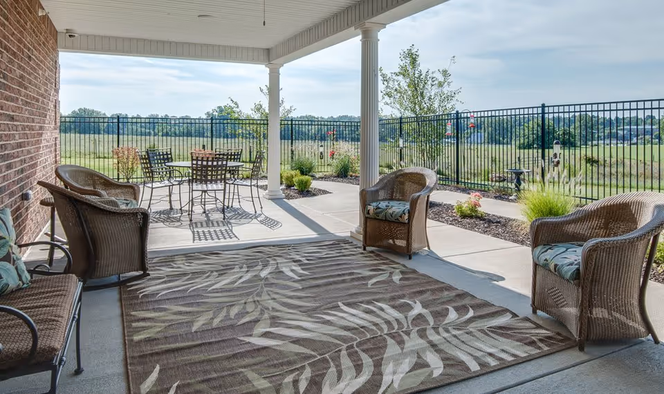 Covered outdoor patio with wicker chairs, a patterned rug, and a metal dining set overlooking a fenced grassy landscape.
