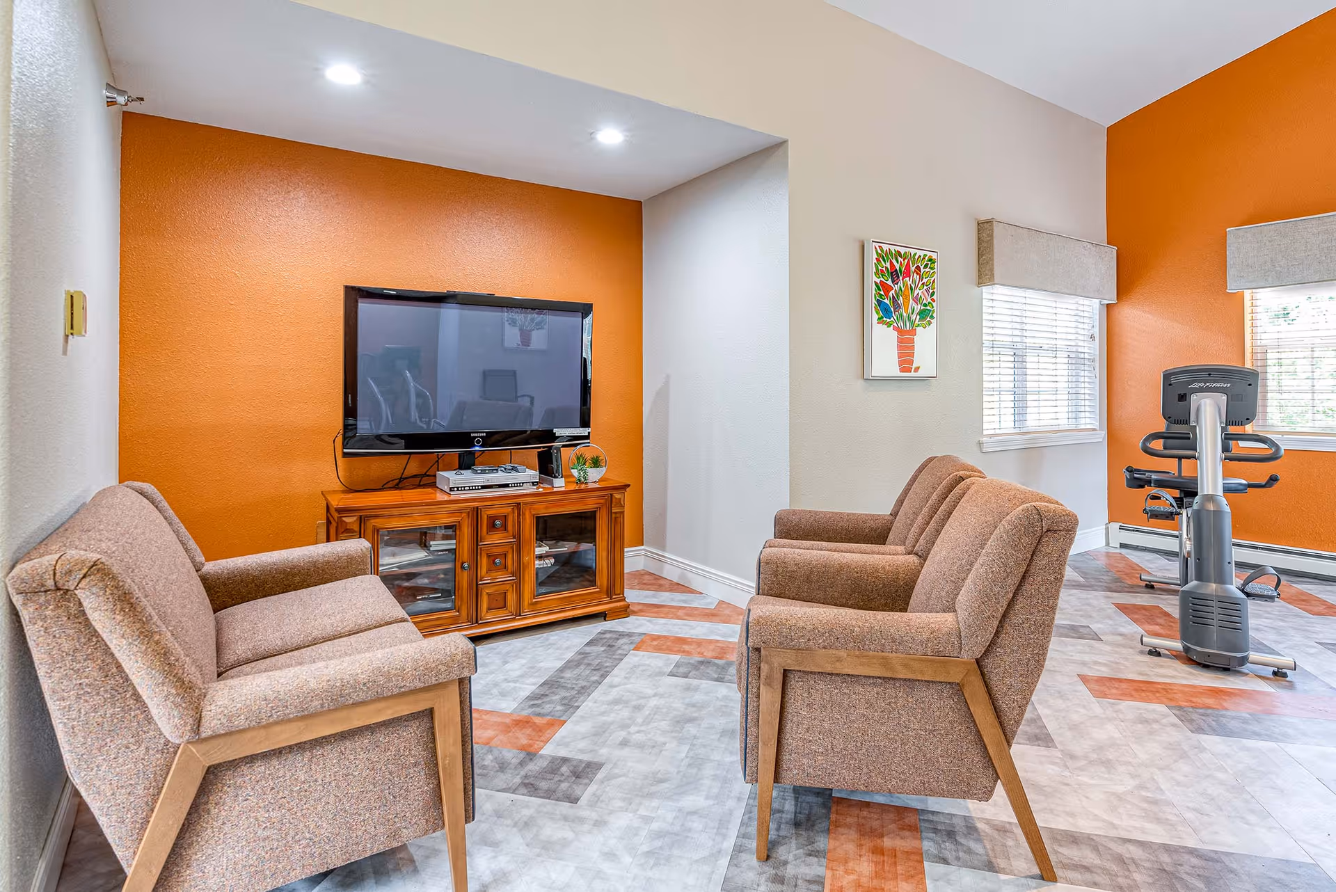 A cozy senior living common area with four cushioned chairs arranged facing a flat-screen TV mounted on an orange accent wall. The TV sits on a wooden cabinet with glass doors. The room has patterned flooring with shades of gray and orange, two windows with blinds, a colorful framed artwork on the wall, and an exercise bike near the windows.