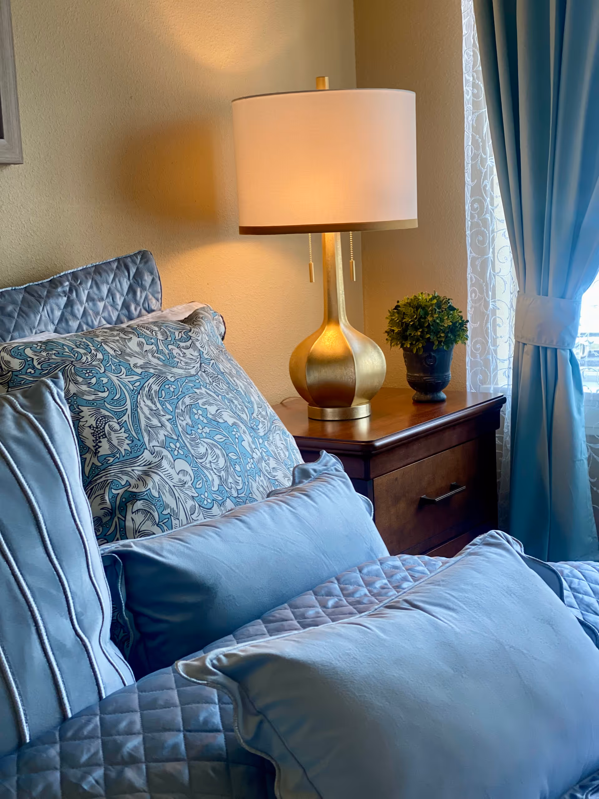 Close-up view of a neatly made bed with multiple blue and patterned pillows, a wooden nightstand with a gold table lamp and a small potted plant, next to a window with sheer white and blue curtains.