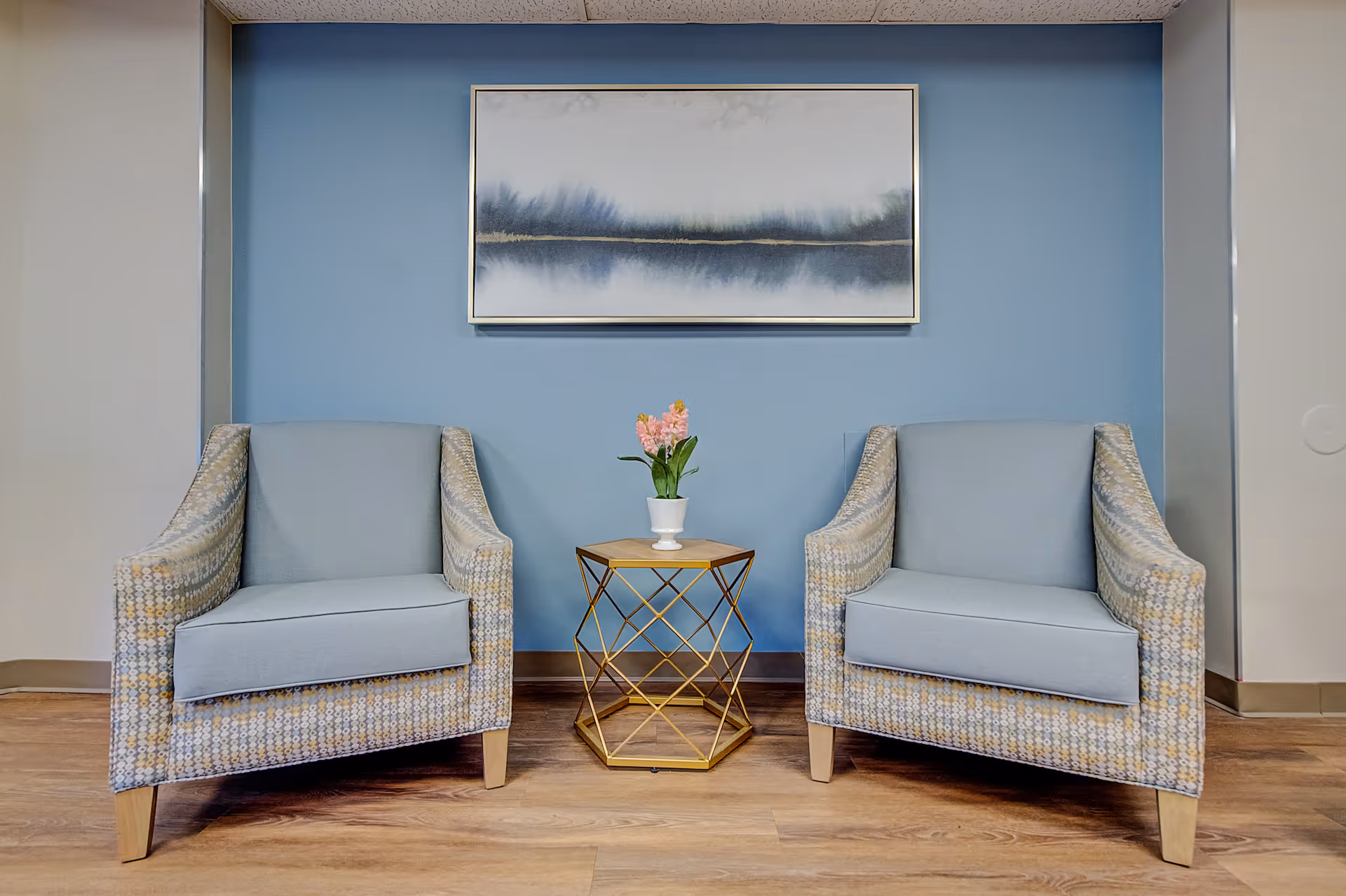 Two patterned armchairs with light blue cushions are placed on either side of a small gold geometric side table holding a potted pink flower. Behind the chairs is a blue wall with a framed abstract landscape painting hanging above.