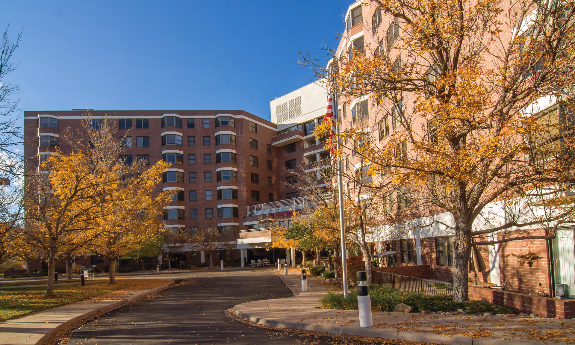 Exterior view of a multi-story senior living facility building with red brick walls, surrounded by trees with autumn foliage under a clear blue sky. A driveway and sidewalk lead to the entrance, with an American flag on a pole near the building.