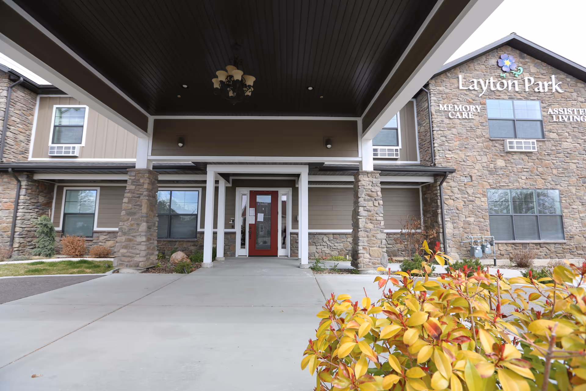 The covered entrance and porte-cochère of the Layton Park Memory Care building with a stone facade, a red front door, and shrubs in the foreground.