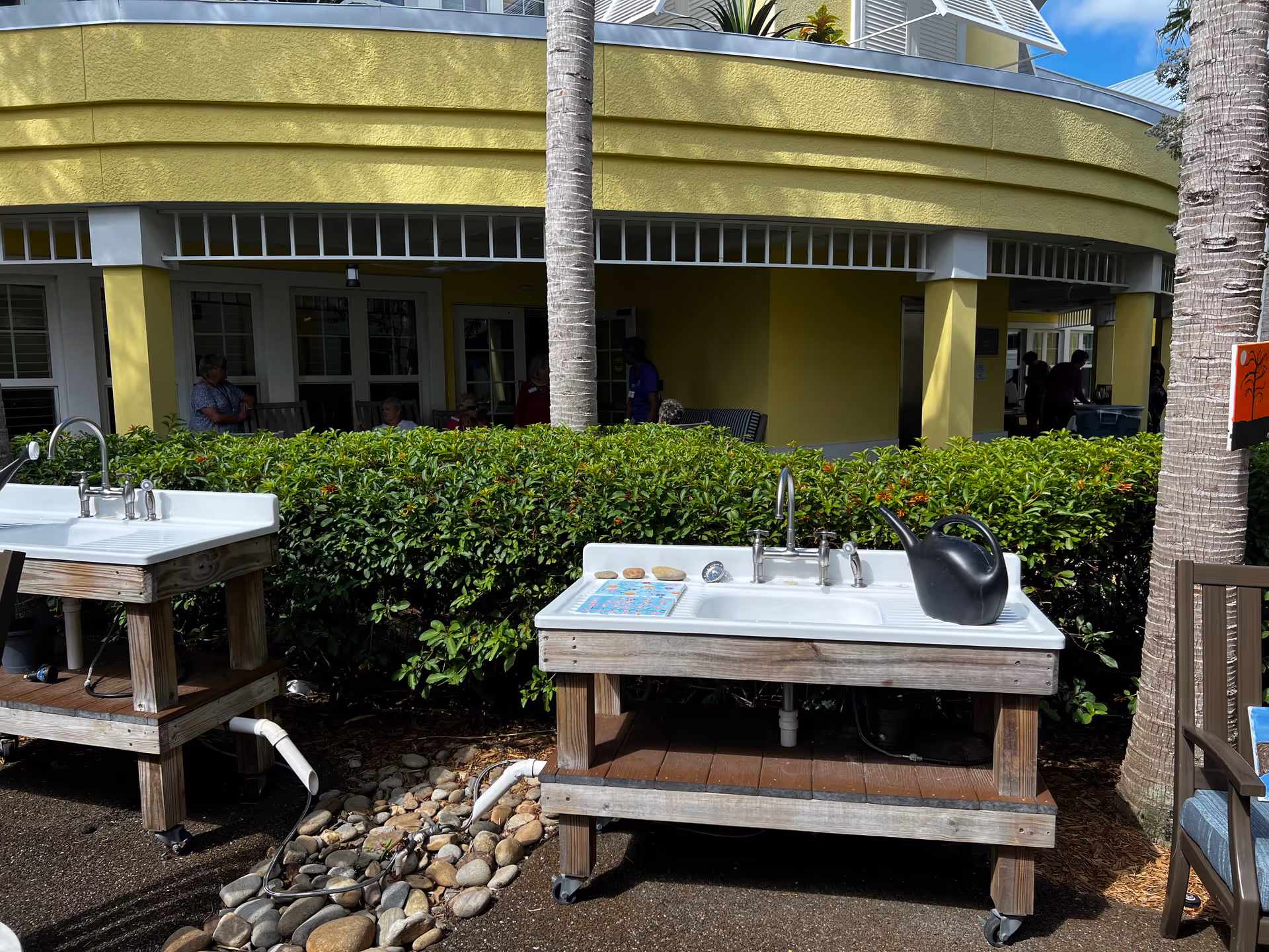 Outdoor area with two white sinks mounted on wooden stands, surrounded by greenery and palm trees. In the background, there is a yellow building with windows and people sitting under a covered patio.