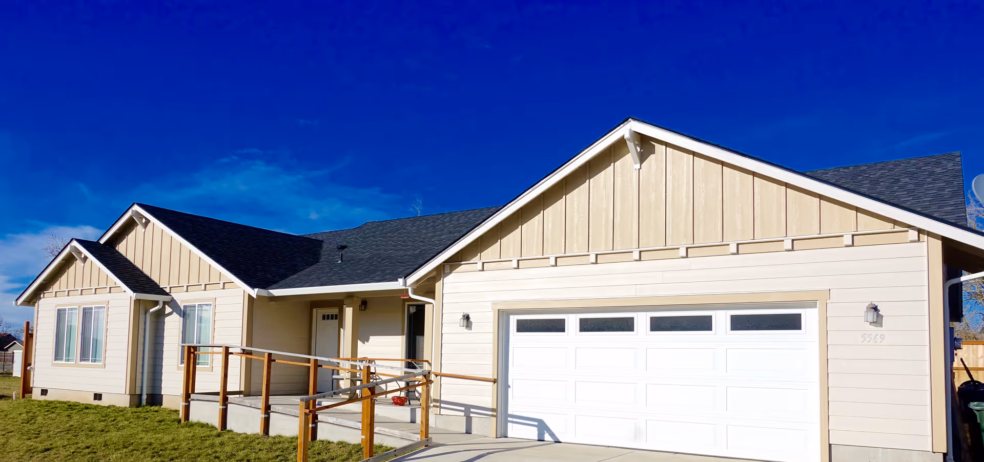 Exterior view of a single-story beige house with a dark roof under a clear blue sky. The house features a white garage door, several windows, a front door, and a wooden ramp with handrails leading to the entrance. The lawn in front is green and well-maintained.