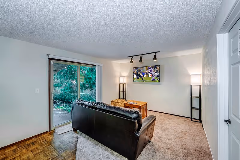 A small living room with a black leather couch facing a wall-mounted flat screen TV showing a football game. The room has beige carpet, two tall floor lamps with white shades on either side of the TV, a wooden coffee table, and a sliding glass door leading outside with greenery visible.