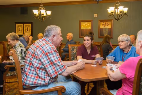 A group of elderly people and a caregiver sitting around tables in a well-lit room playing card games. The room has green walls, framed floral artwork, and chandeliers hanging from the ceiling.