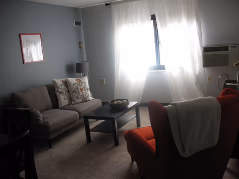 A small living room with a gray loveseat, a red armchair draped with a blanket, a black coffee table, and a window with sheer white curtains.