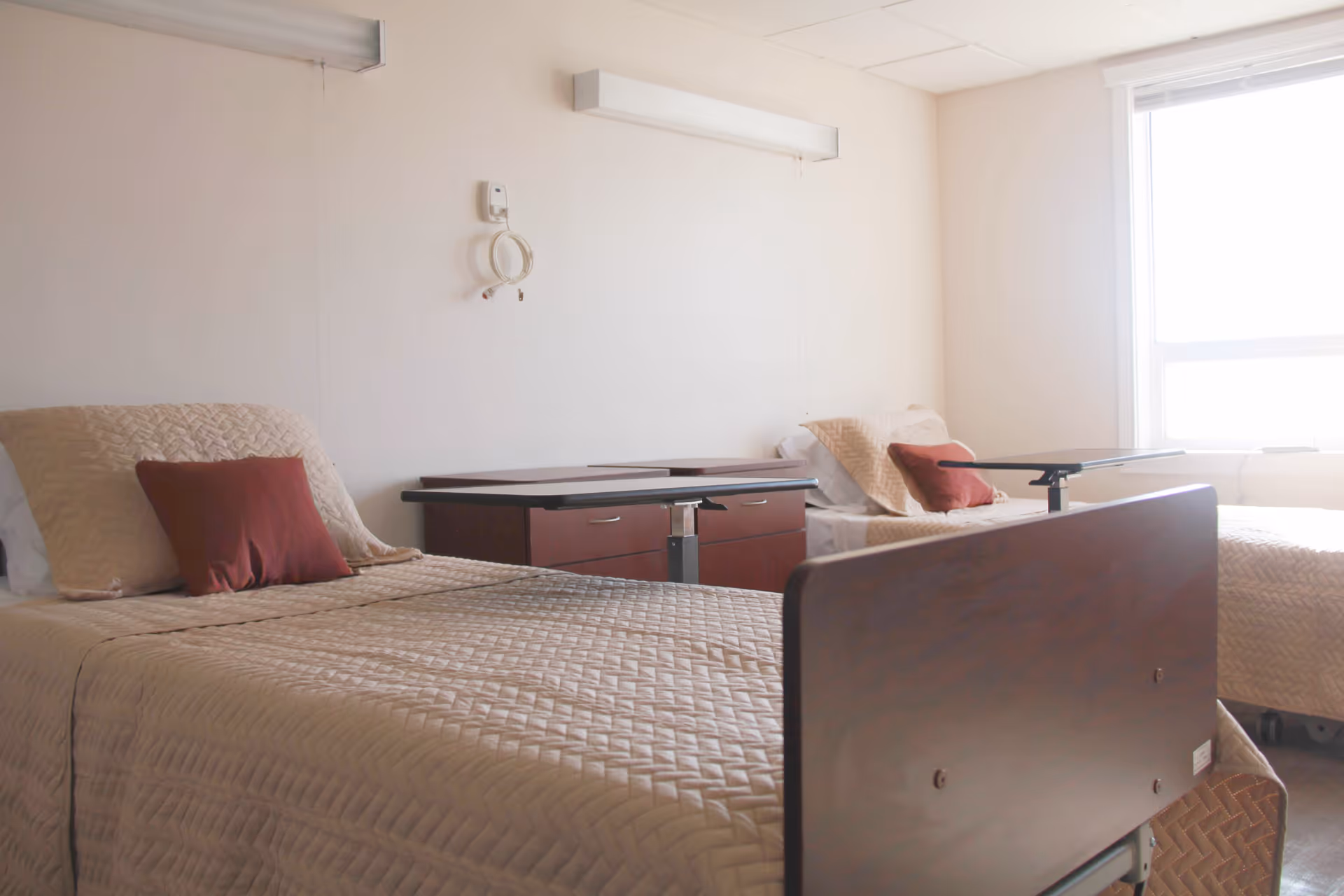 A bright room with two hospital-style beds, each with beige quilted bedspreads and red pillows. Between the beds are two overbed tables and a wooden cabinet. A large window on the right side lets in natural light, and the walls are plain white with two wall-mounted light fixtures above the beds.