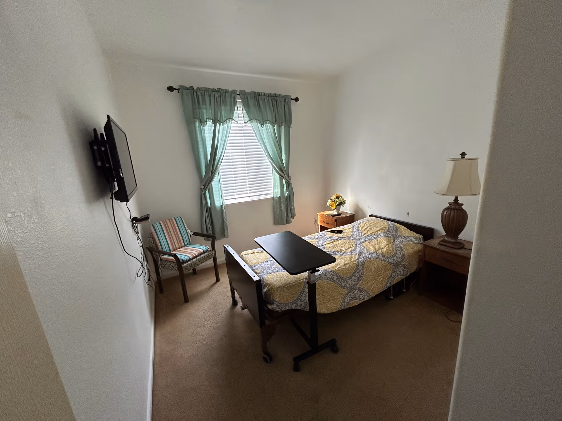 Small private bedroom with a single bed covered by a yellow patterned comforter, an overbed table, nightstands with a lamp and flowers, a striped chair, wall-mounted TV and a window with green curtains.